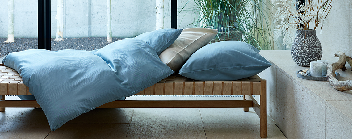 Ropa de cama de color gris sobre edredón y almohada cubriendo un sofá cama frente a una ventana. 