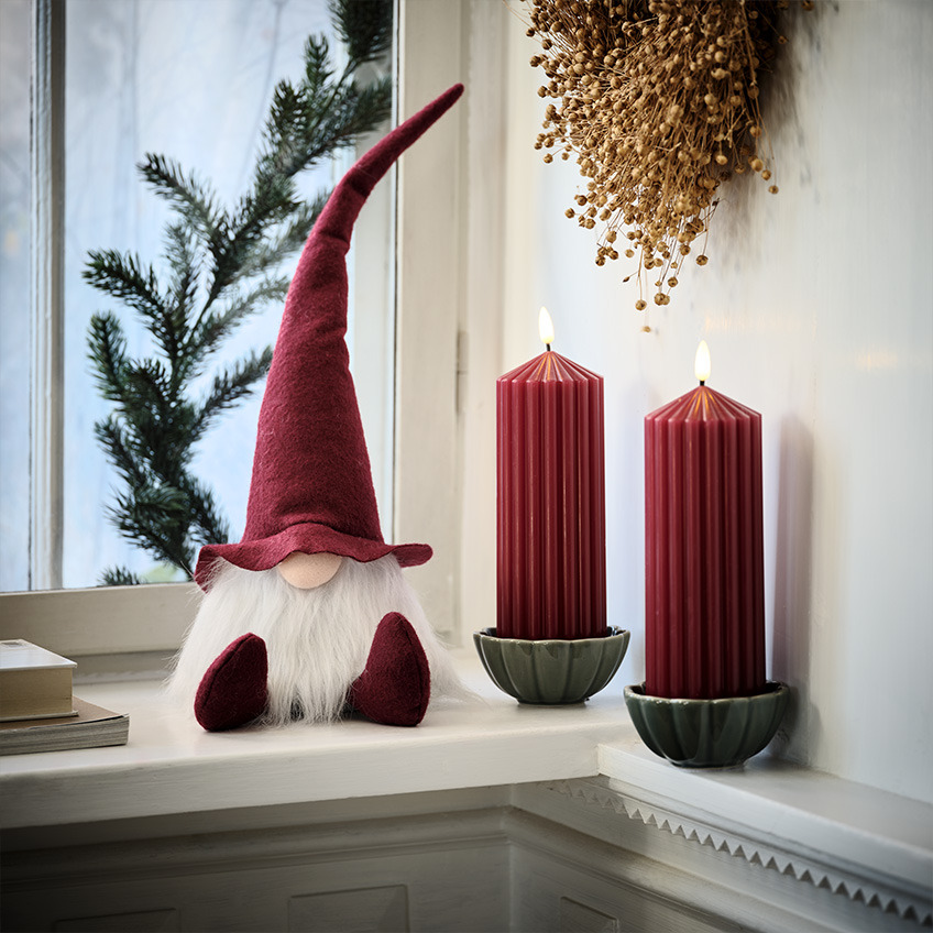 Duende navideño escandinavo en la repisa de la ventana junto a velas rojas grandes.