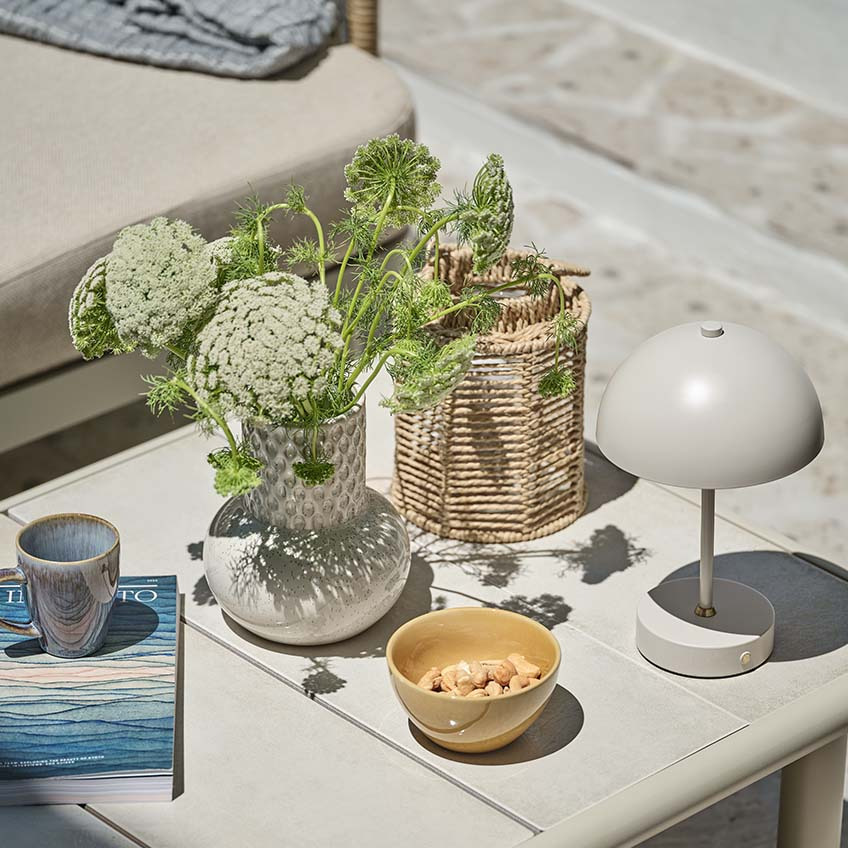 Pequeña mesa de jardín con varias decoraciones, incluyendo una lámpara a pilas pequeña, un jarrón y un farol