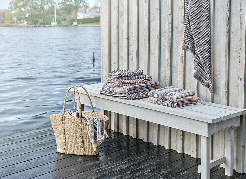 IDRE towels in grey or sand and SEGERSTA striped towels Luxury towels on a bench and in a wicker bag by a lake