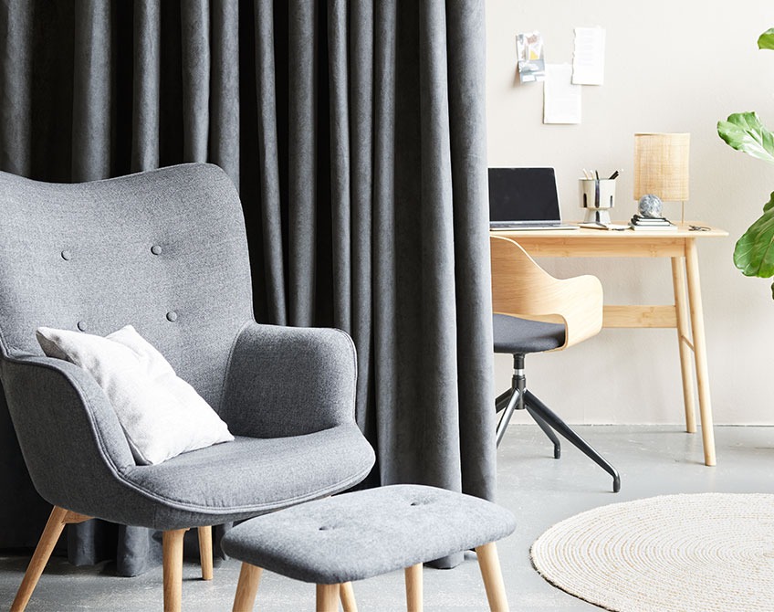 Grey curtains separating a home office with desk and office chair from a living room with an armchair and footstool