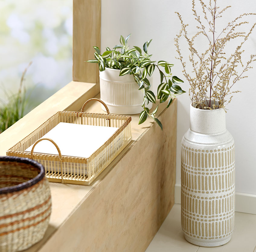 Jarrón alto junto al alféizar de la ventana con bandeja de bambú y maceta blanca con planta artificial